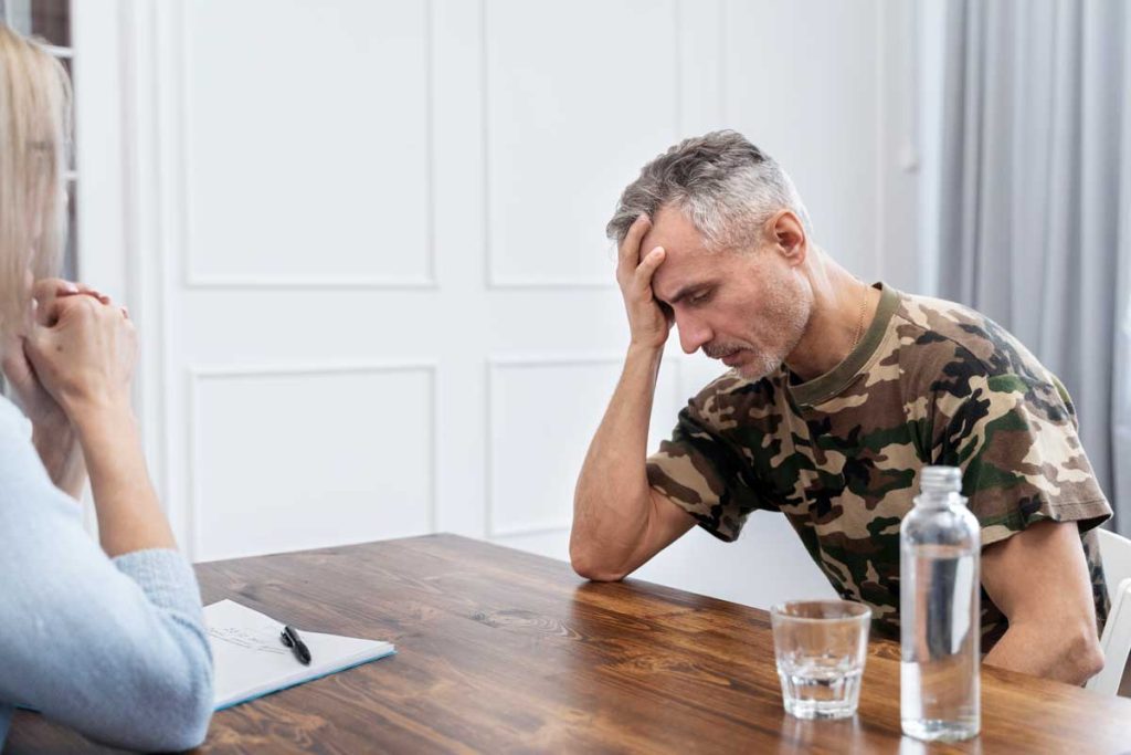 man wearing a camp print shirt sitting at a table with his hand on his forehead experiencing the pain of withdrawal symptoms