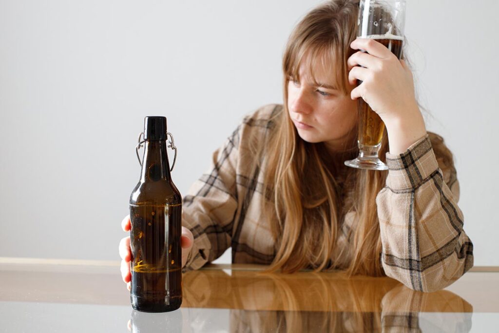 Woman in distress with a beer glass held to her hand and a bottle in the other hand sitting at a table dictating the need for professional support for alcoholism