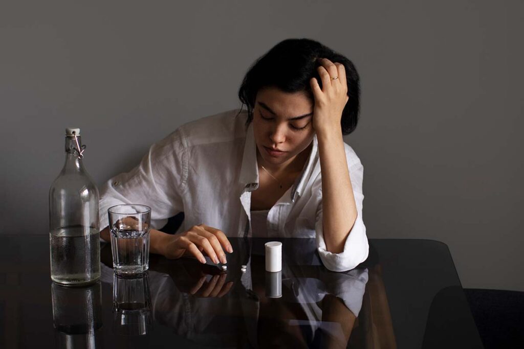 Woman in distress sitting at a table with pills and a glass of water, symbolizing emotional pain and substance dependence—Capistrano Beach Recovery offers compassionate care for those struggling with addiction and mental health challenges.